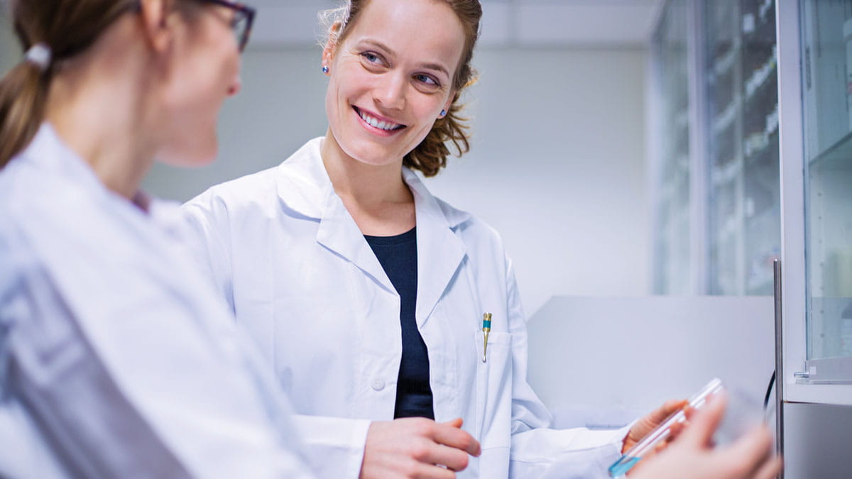 Enzymes, NGS, Two female scientists both Caucasian discussing results one holing a test-tube with blue liquid in a laboratory setting